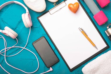 Evde fitness - bir kağıt yaprak kalem, dambıl ve beyaz spor ayakkabı, bir havlu ve kulaklık dokulu mavi bir arka planda cep telefonu