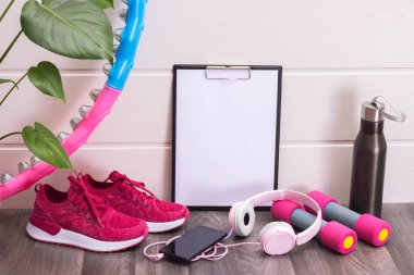 Pembe spor ayakkabılar, dambıllar, tahta, kulaklık ve cep telefonu, yeşil büyük yapraklar ve beyaz arka planda bir şişe su.