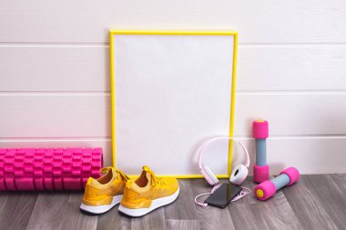 Masaj silindiri ve dambıllar, sarı spor ayakkabılar, tahta, kulaklık ve beyaz arka planda cep telefonu.