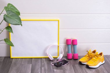 Tahta, halterler, sarı spor ayakkabılar, kulaklıklar ve bir cep telefonu ve beyaz arka planda büyük yeşil yapraklar.