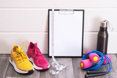 Pembe ve sarı spor ayakkabılar, dambıllar ve not panosu, mezura ve su şişesi, yakın plan.
