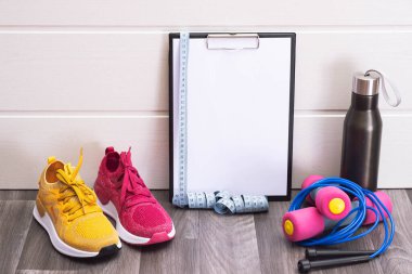 Spor malzemeleri - pembe ve sarı spor ayakkabılar, dambıllar ve bir not panosu, mezura ve su şişesi, yakın çekim
