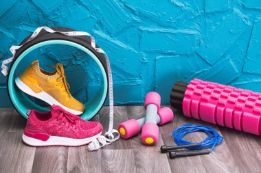Evde egzersiz - sarı ve pembe spor ayakkabılar, dambıllar, atlama ipi, ölçü bandı ve masaj ruloları, mavi arka planda.
