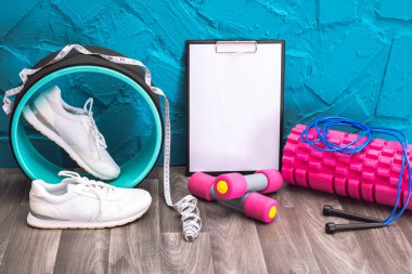 Evde egzersiz - beyaz spor ayakkabıları, dambıllar, ip atlama, mezura, plaka ve masaj silindiri, mavi arka planda.