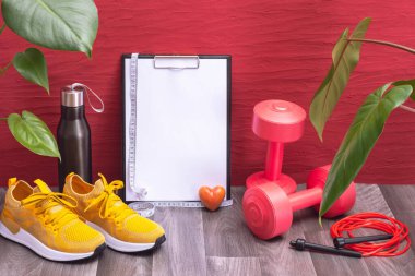 Evde egzersiz - kırmızı halterler, su şişesi ve ip atlama tahtası ve ölçüm bandı, sarı spor ayakkabılar ve büyük yeşil yapraklar ve küçük kalp, kırmızı arka plan