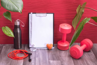 kırmızı halterler, su şişesi ve ip atlama tahtası ve ölçüm bandı, kırmızı arka planda büyük yeşil yapraklar ve küçük kalp.