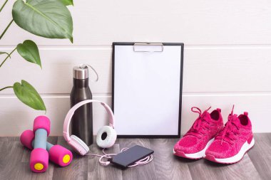 Pembe spor ayakkabılar, dambıllar, su şişesi ve not panosu, kulaklıklar ve cep telefonu, yeşil büyük yapraklar ve beyaz arka plan