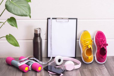 çok renkli spor ayakkabılar, dambıllar, su şişesi ve not panosu, kulaklık ve cep telefonu, yeşil büyük yapraklar, beyaz arka planda.
