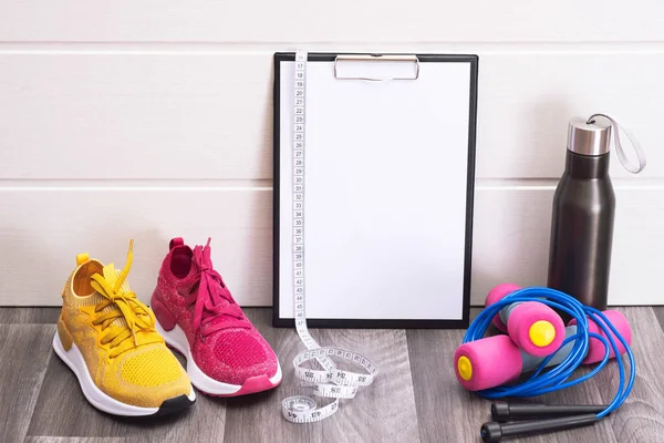 Pembe ve sarı spor ayakkabılar, dambıllar ve not panosu, mezura ve su şişesi, yakın plan.