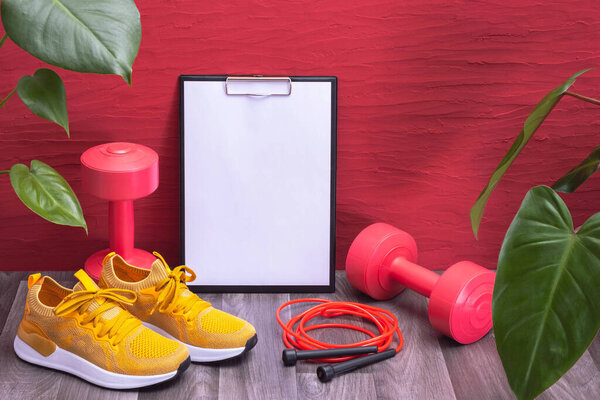 home workout - red dumbbells, jump rope and yellow sneakers, note board and green large leaves on a red background, close-up