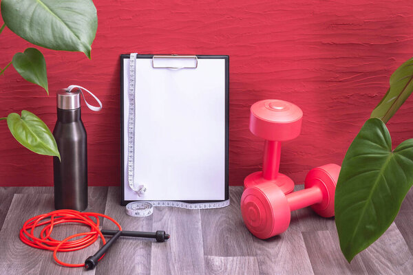 workout at home - red dumbbells, water bottle and jump rope, note board and measurement tape, large green leaves