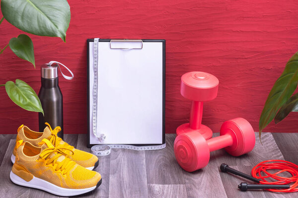 workout at home - red dumbbells, water bottle and jump rope, note board and measurement tape, yellow sneakers and big green leaves, red background
