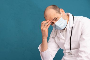 Maskeli yorgun bir doktor, elini alnına dayadı, mavi bir arka planda.
