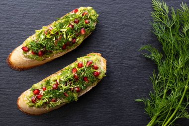 İki aperatif - avokado, olgun nar tohumları serpiştirilmiş, susam tohumları ve mis kokulu taze dereotu, siyah arka planda ve yakın planda uzanır.