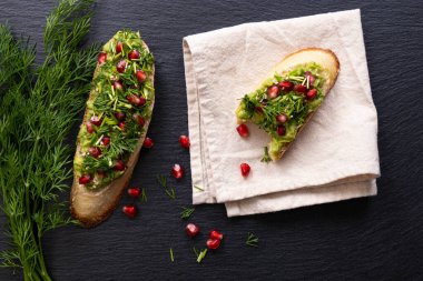 Bir peçeteye ısırılmış avokado, nar ve dereotu tohumları, biri ısırılmış, taze dereotu dalları ve başka bir aperatif, siyah arka plan.