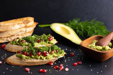 Avokado kanapeleri ve olgun nar tohumları, susam tohumları ve aromatik dereotlar, siyah arka planda.