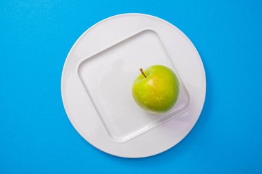 Mavi arka planda yeşil elmalı beyaz bir plaka duruyor, yakın plan.