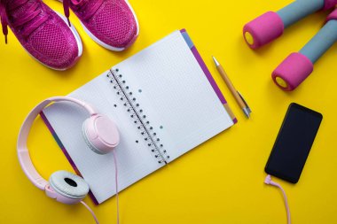 Pembe spor ayakkabılar, dambıllar, sarı arka planda cep telefonlu kalem ve kulaklıklar.