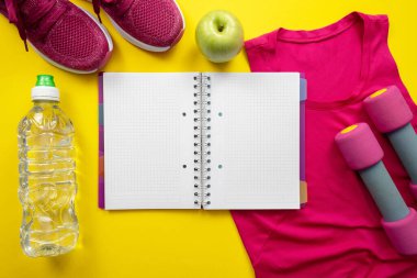 Pembe tişört, spor ayakkabı ve dambıllar, not defteri, elma ve su şişesi sarı arka planda.