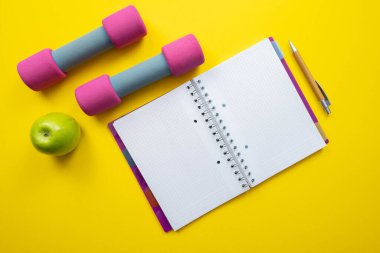 Pembe halterler, defter, yeşil elma ve sarı arka plan.
