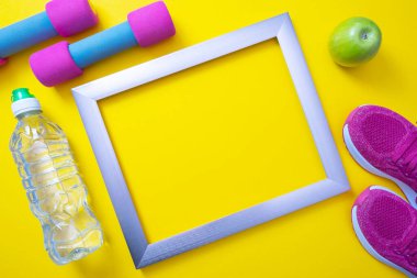 Metal çerçeve, yeşil elma ve bir şişe su, pembe spor ayakkabılar ve halterler sarı arka planda uzanır.