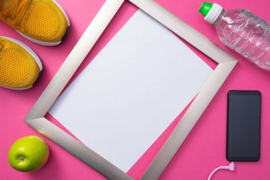 Beyaz arka planlı metal çerçeve, cep telefonu ve sarı spor ayakkabılar, bir şişe su ve genel pembe arka planda yeşil elma.