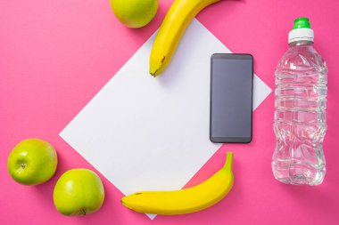 Vitaminler - elmalar ve muzlar, bir cep telefonu ve bir şişe su, ortak pembe arka planda uzanıyorlar.