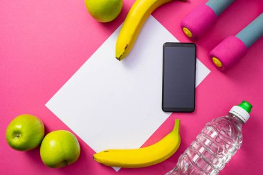bir cep telefonu ve bir şişe su, dambıllar, elmalar ve muzlar ortak pembe arka planda uzanır.