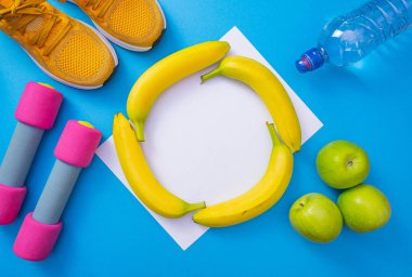 Beyaz kağıt, dört muz, elmaların, garnitürlerin, suyun ve spor ayakkabıların yanında, ortak bir mavi arka planda yer alır.