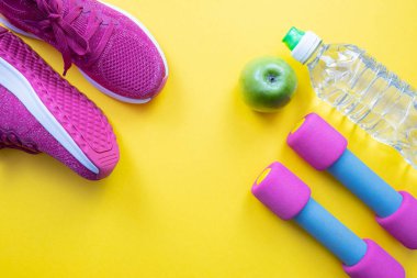 Pembe spor ayakkabılar ve dambıllar, yeşil elma ve bir şişe su sarı arka planda, yakın plan