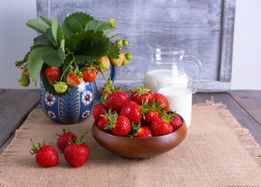 Seramik bir vazoda çilek ve ahşap bir kasede çilek ve bir sürahi süt.