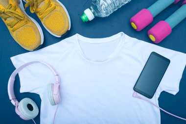 Beyaz tişört ve sarı spor ayakkabılar, bir şişe su ve dambıl, kulaklıklı cep telefonu, mavi arka plan.