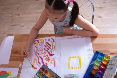 Kız masaya oturur ve bir fırça ve boyalarla resim çizer, üstten manzara