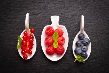 olgun böğürtlen - ahududu, kırmızı frenk üzümü ve yaban mersini ve nane yaprağı, siyah arka planda beyaz bir tabakta uzanır.