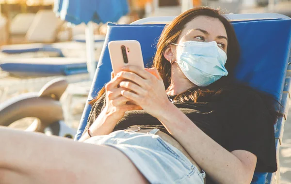 Kobieta w medycznej masce chirurgicznej siedzi na publicznej plaży i za pomocą smartfona — Zdjęcie stockowe
