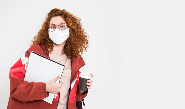Portret pięknej rudej studentki w masce medycznej trzymającej zeszyty ćwiczeń. badanie w czasie pandemii koronawirusa covid 19 — Zdjęcie stockowe