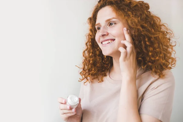 Krásná usmívající se žena s červeným afro kadeřnictví leapplying kosmetické krém léčba na její obličej. — Stock fotografie