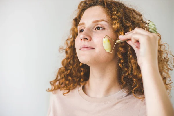 Beautiful woman with red afro hairsty leapplying cosmetic cream treatment on her face with roller massage tool — Stock Photo, Image