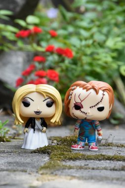 Korku filmlerinden Chucky ve Tiffany 'nin seri katil bebeklerinin aksiyon figürleri. Yaz bahçesinde kırmızı güller ve yosunlu asfalt levhalar.