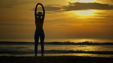 Spor kıyafetli bir kadın plajda gün batımında egzersiz yapıyor..