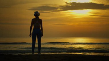 Gün batımında plajda yoga yapan genç bir kadının silueti, sağlıklı yaşam tarzı ve spor konsepti..