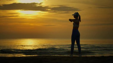 Gün batımında sahilde mayo giymiş, sörf tahtası olan bir kadının silueti..