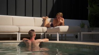 Mayolu ve güneş gözlüklü bir adam havuz kenarında dinleniyor.