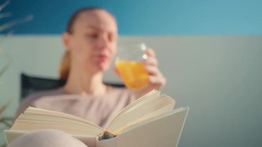 Açık kahverengi saçlı bir kadın, açık havada bir sandalyede otururken, açık mavi bir gökyüzü ve beyaz bir duvarla, etrafında barışçıl bir atmosfer yaratarak kitap okumaktan hoşlanıyor.