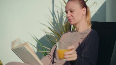 Açık havada bir sandalyede oturan kadın elinde bir bardak portakal suyu, yeşil bitkiler ve arka planda berrak mavi bir gökyüzü olan bir kitap okuyarak sakin bir atmosfer yaratıyor.