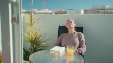 Kadın rahat rahat bir şekilde balkonda oturmuş meyve suyunu yudumluyor ve kitap okuyor, canlı bitkiler ve açık havada sakin bir hava yaratıyor.