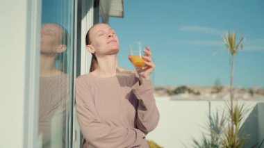 Uzun sarı saçlı bir kadın, açık havada durur, kitap okur ve sayfalarını çevirir, yanında yeşil bir bitki, açık mavi gökyüzüne karşı parlak, güneşli bir ortamda görünür.