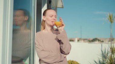 Uzun sarı saçlı bir kadın, açık havada durur, kitap okur ve sayfalarını çevirir, yanında yeşil bir bitki, açık mavi gökyüzüne karşı parlak, güneşli bir ortamda görünür.