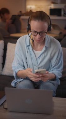 Açık mavi gömlekli ve kulaklı beyaz bir kadın, arka planda rahat bir oturma odası olan akıllı telefon ve dizüstü bilgisayar kullanarak koltukta otururken video görüşmesi sırasında etkileşime giriyor.