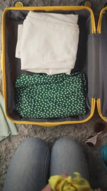 Açık mavi gömlekli kadın sarı eşarp ve hasır şapkayı içinde giysi, kulaklık ve seyahat aksesuarları bulunan yumuşak, desenli bir yüzeye koymaya devam ediyor.